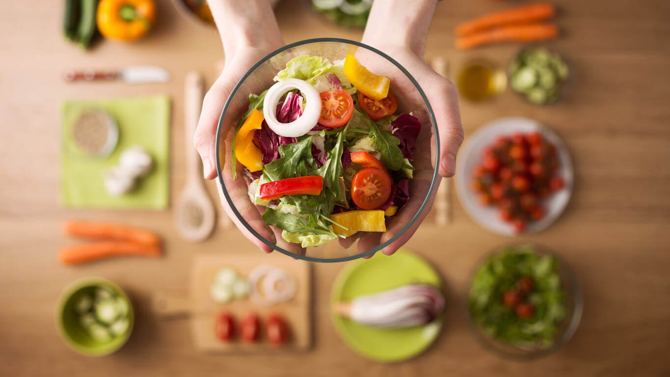 Hands holding an healthy fresh vegetarian salad in a bowl, fresh raw vegetables on background, top view.